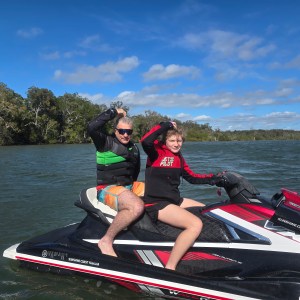 Two people on a jet ski waving on a sunny day with trees and water in the background.